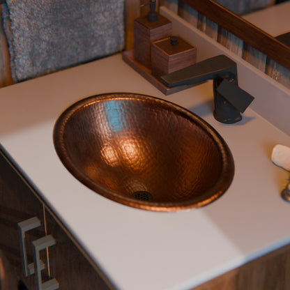 Oval rolled rim copper drop-in bathroom sink in antique finish, traditional style by Novatto, shown in a dark matte setting
