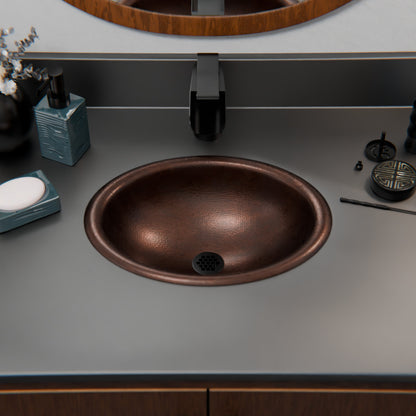 Small oval copper drop-in bathroom sink in antique finish, transitional style by Novatto, with dark interior and matching drain assembly.