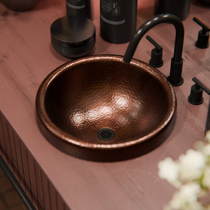 Round copper drop-in bathroom sink by Novatto, traditional style with antique finish, shown in a natural setting.