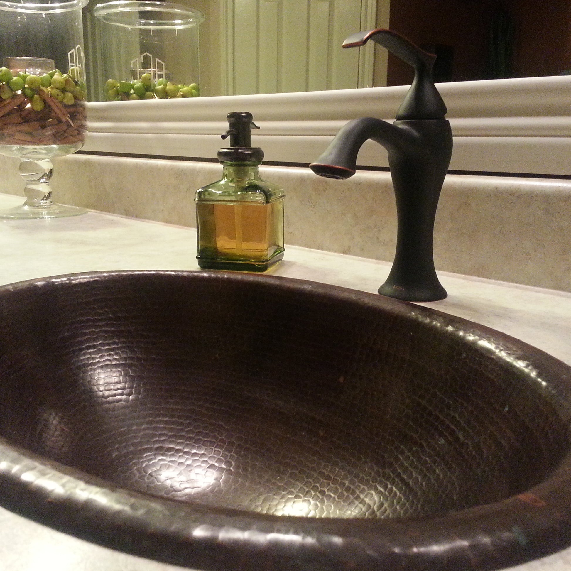 Small oval copper drop-in bathroom sink in antique finish by Novatto, shown installed in a modern transitional bathroom setting.