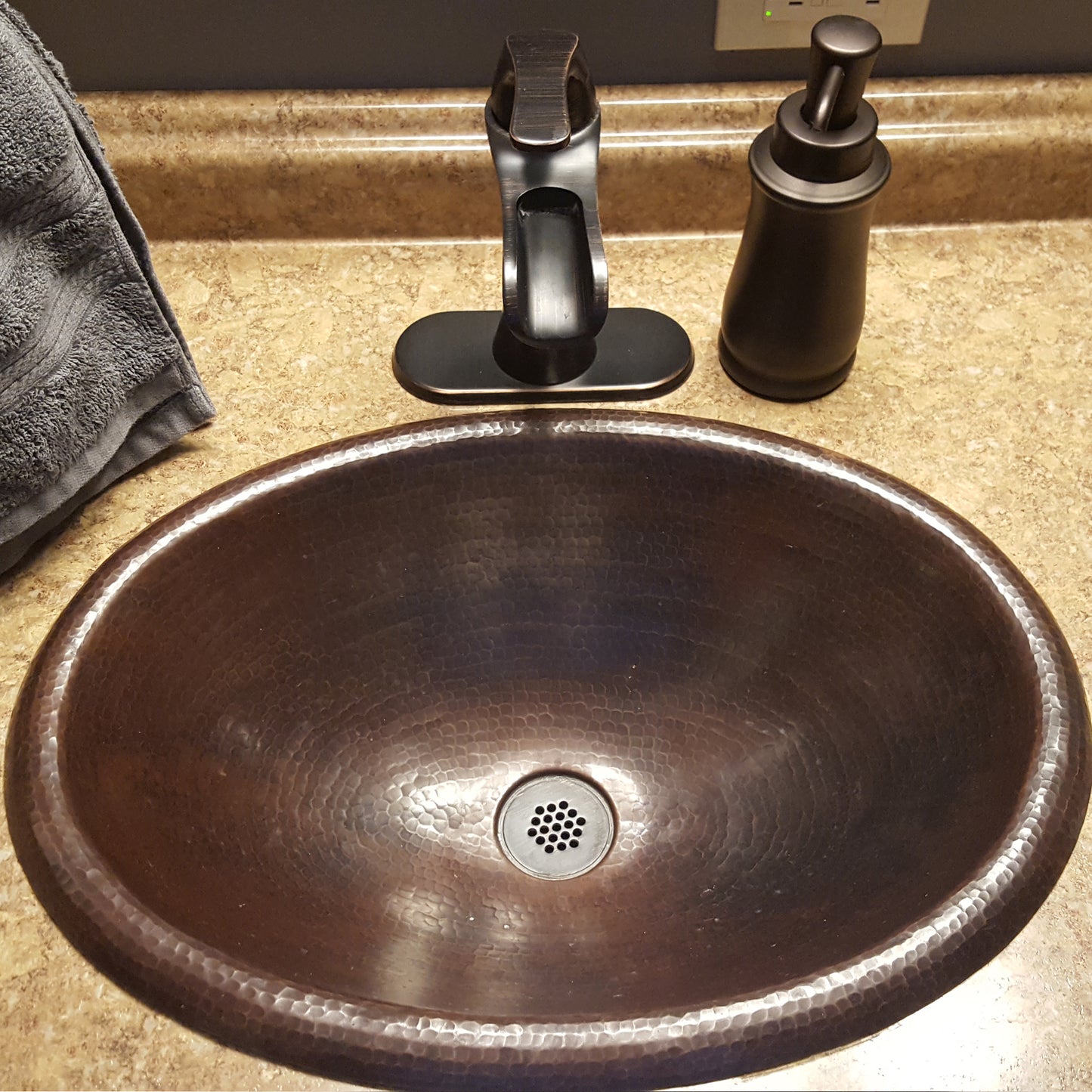 Traditional oval copper drop-in bath sink with rolled rim in antique finish by Novatto, shown in a bathroom setting.