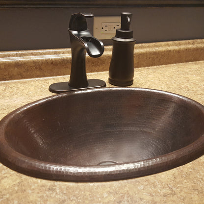 Traditional oval copper drop-in bathroom sink with rolled rim in antique finish by Novatto, shown in a modern bathroom setting.